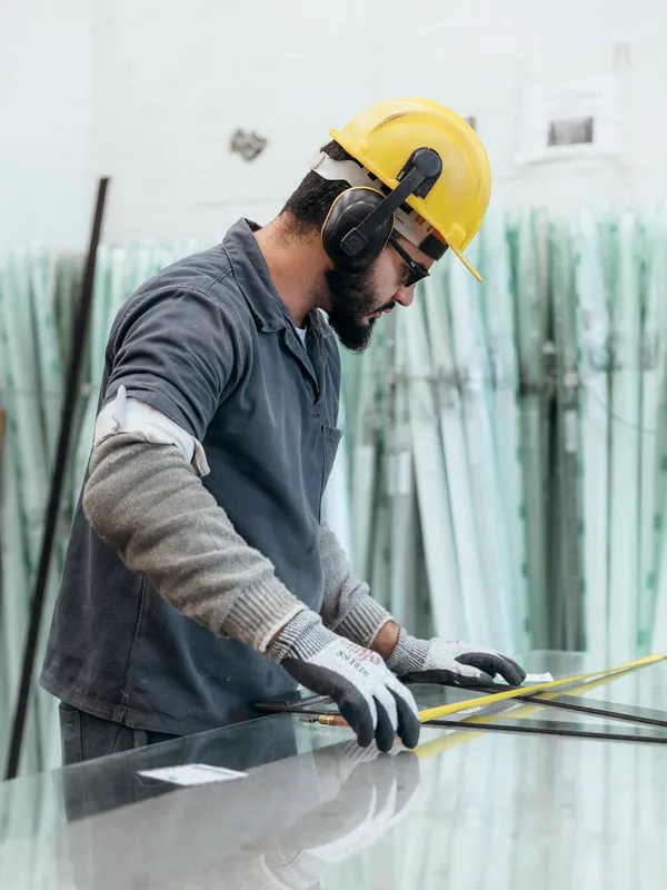 Trabajador con casco y guantes mide una lámina de vidrio en un taller